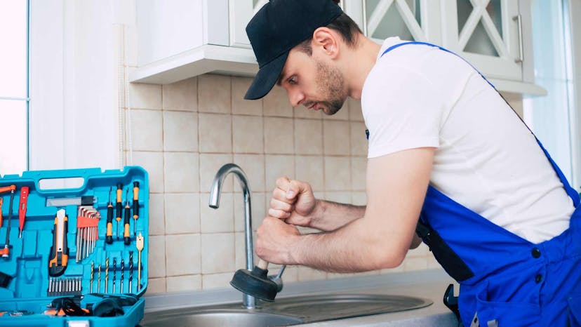 Residential cleaner in blue overalls holding a plunger above the sink with tool box open on counter